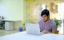Asian man wearing glasses works on laptop in a modern, bright office space.